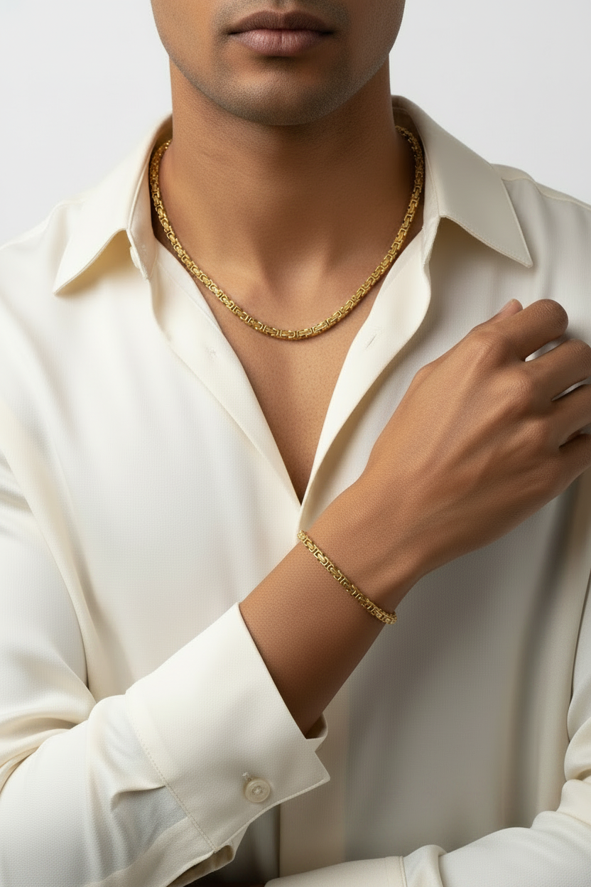 A close-up of a man wearing a gold chain necklace and a matching gold bracelet, styled with a cream-colored collared shirt against a light gray background.
